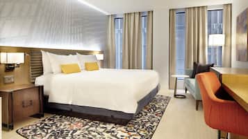 A hotel room with a bed, bedside table, lamp, desk, chair, and a patterned rug.