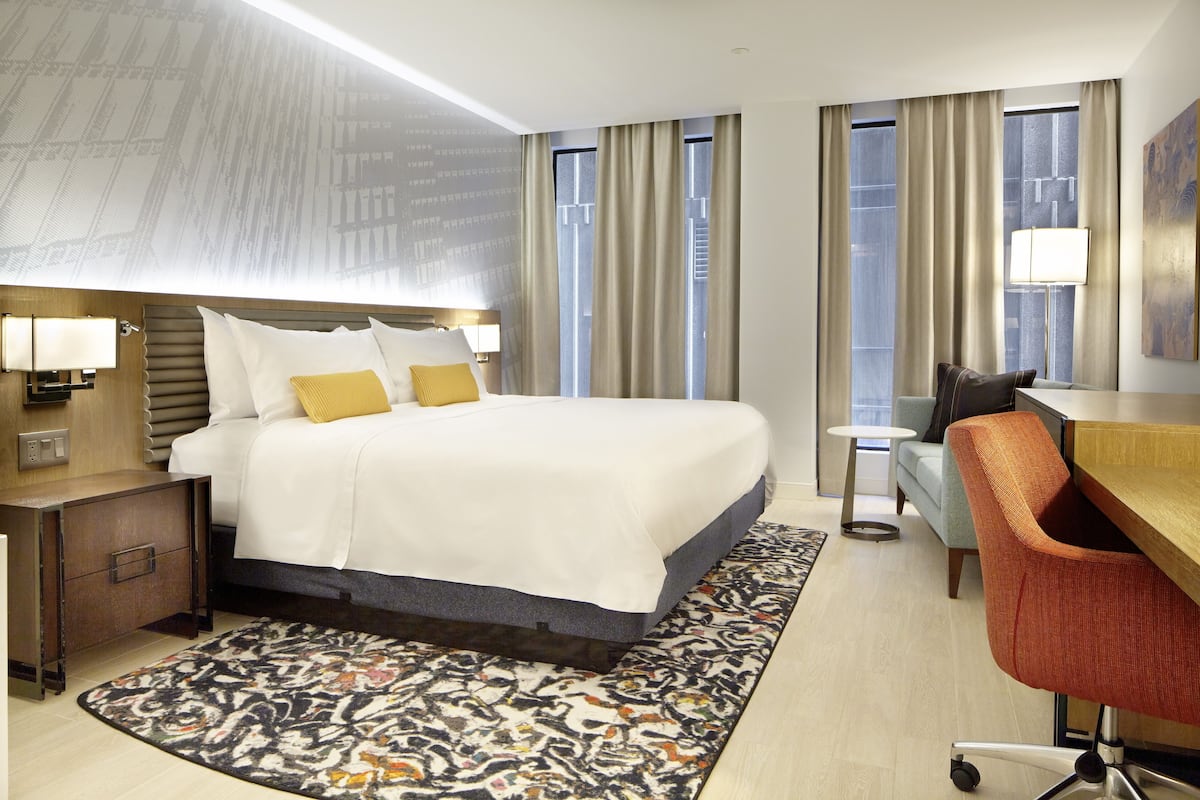 Habitación de hotel con una cama grande, un escritorio, una silla y vista a la ciudad desde las ventanas.