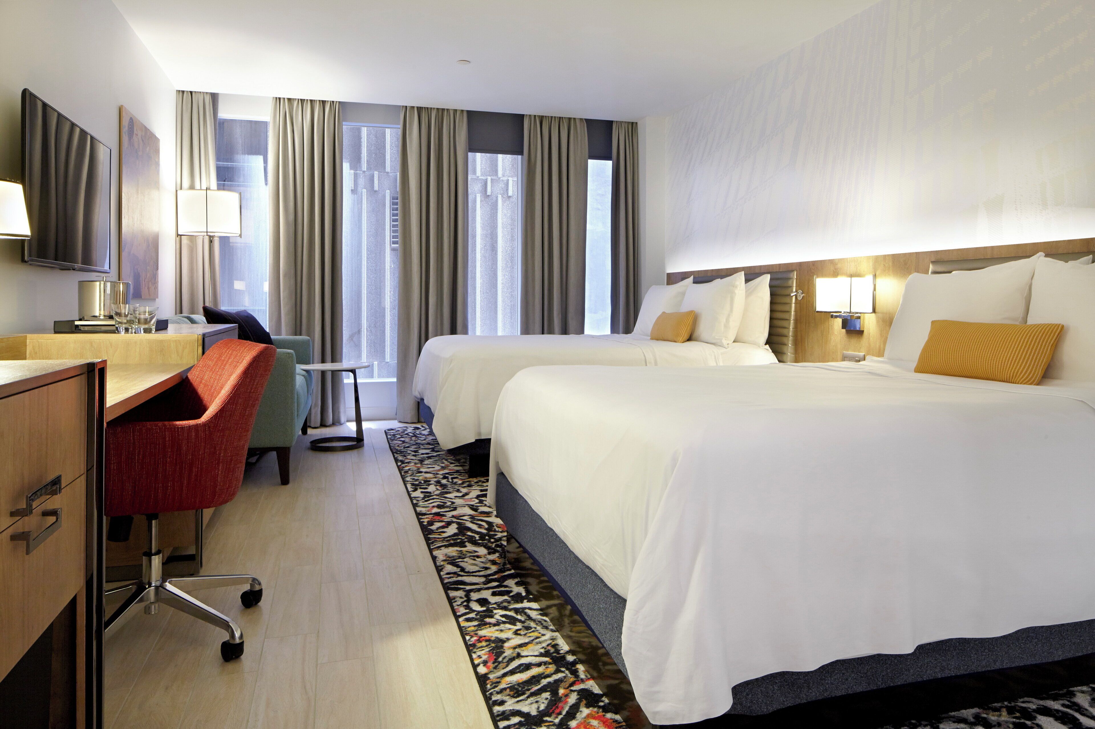 A hotel room with two beds, a desk, a chair, a TV, and a window with curtains.