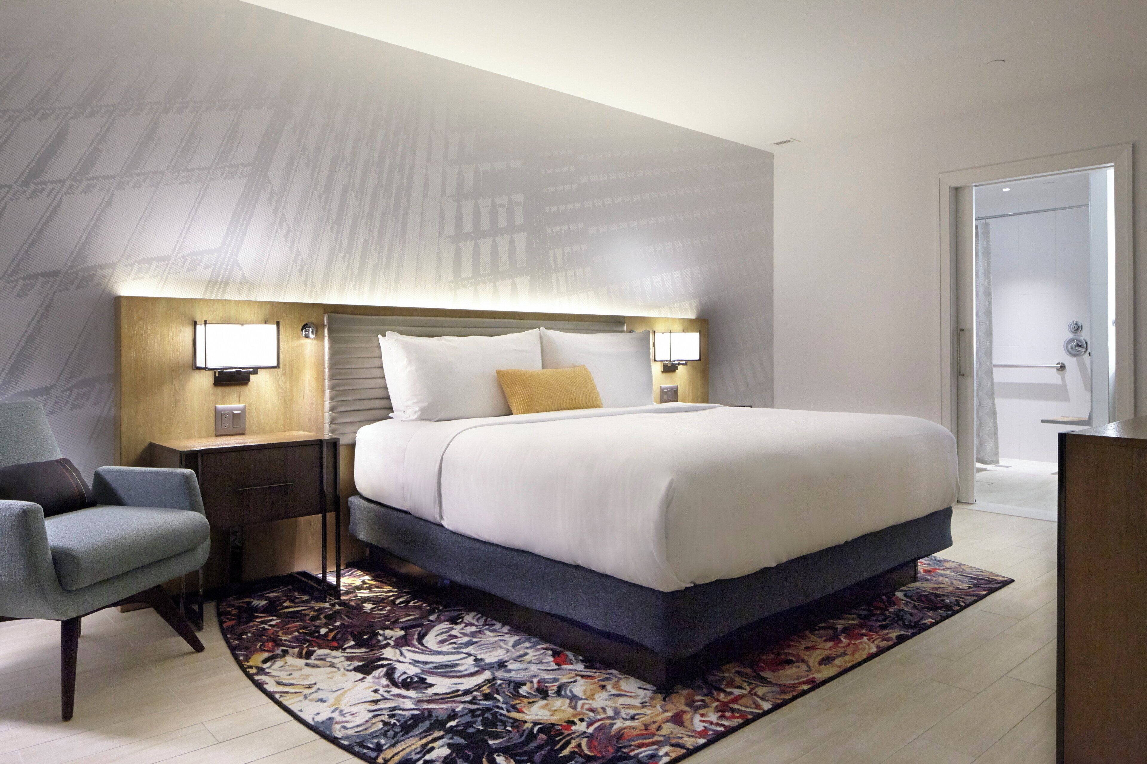 Ein modernes Hotelzimmer mit einem großen Bett, einem Schreibtisch, einem Sessel und einem gemusterten Teppich.