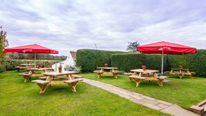 An outdoor patio area with wooden picnic tables and red umbrellas, surrounded by a well-maintained hedge and a stone pathway. - Black Horse Inn  (Northallerton)