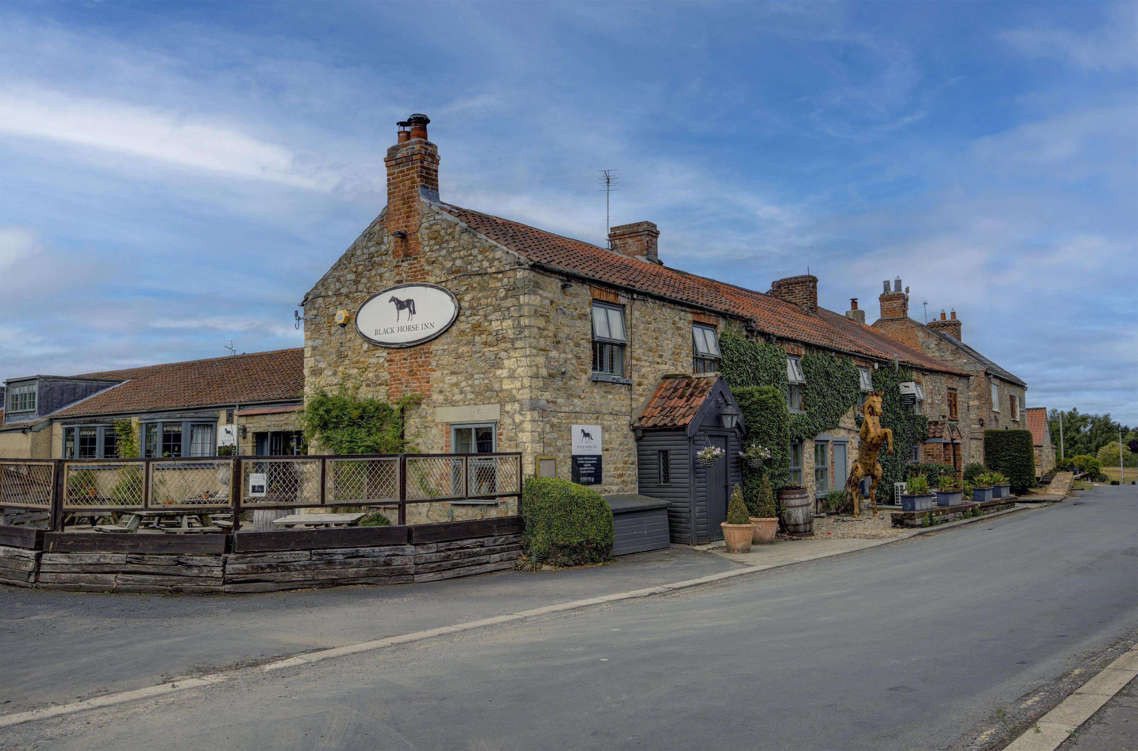 A stone building with a sign for the Black Horse Inn, featuring a traditional brick chimney and a small garden with a horse statue.
