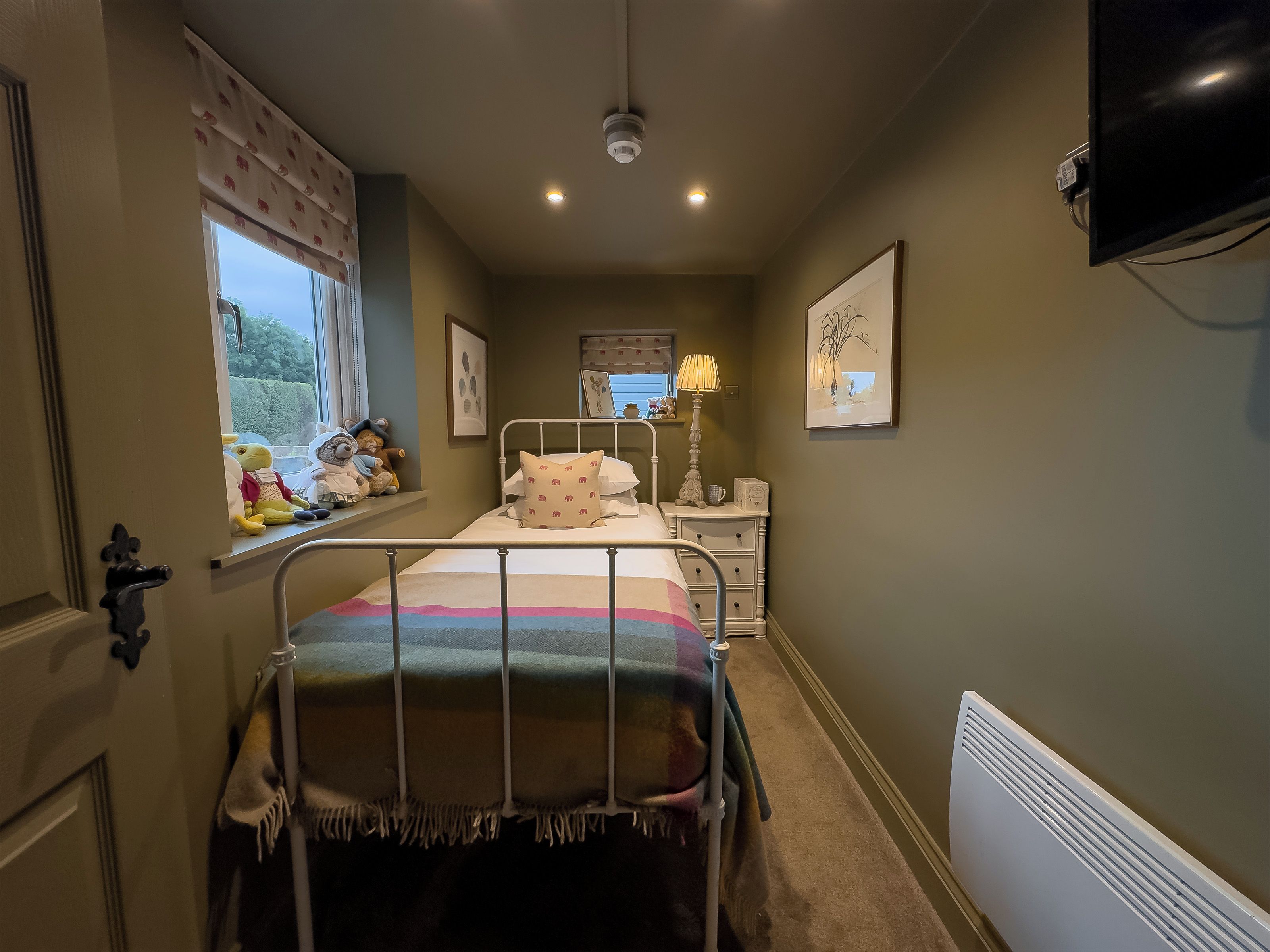 Une chambre à coucher comprenant un lit, une fenêtre, une table de chevet avec une lampe, un radiateur et un téléviseur fixé au mur.