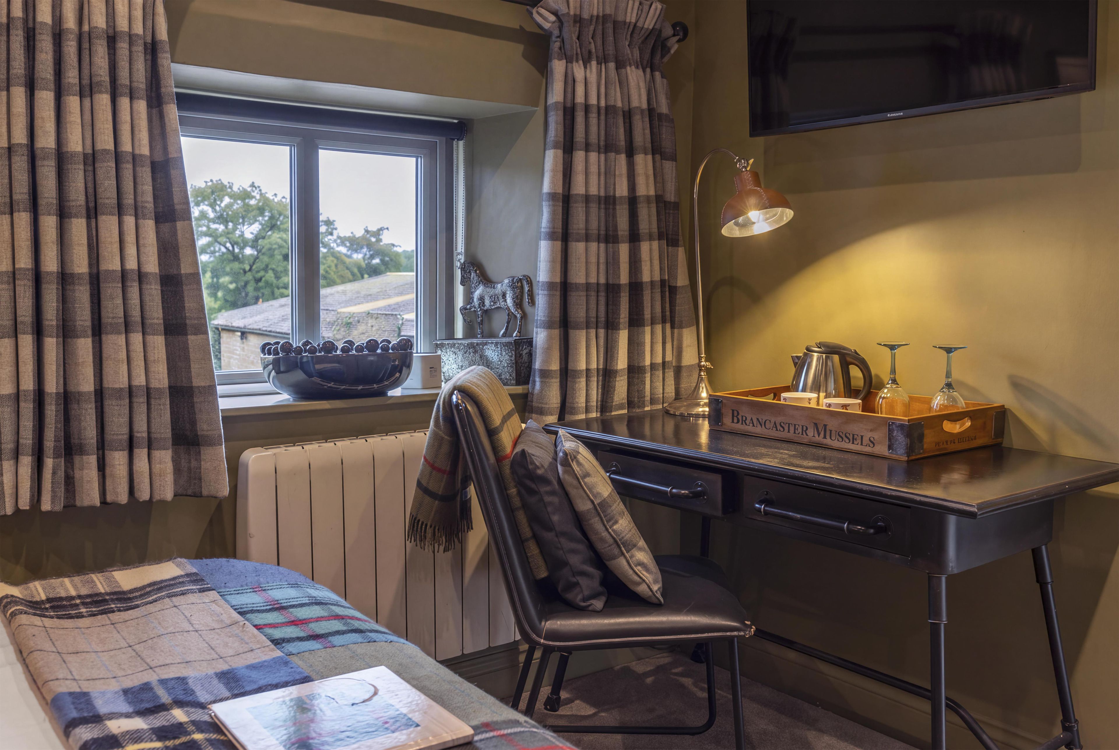 A room with a bed, a desk, a chair, a window with curtains, a radiator, and a small shelf with decorative items.