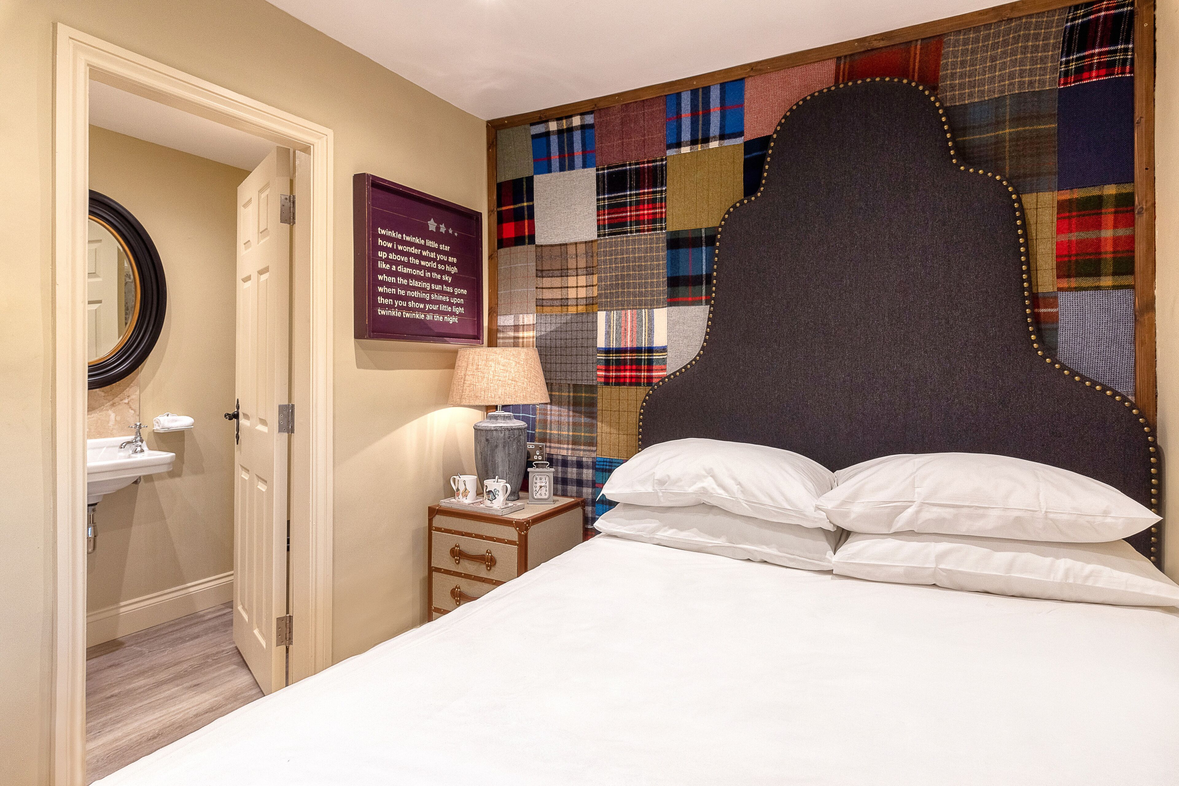 Une chambre avec un grand lit, une table de chevet avec une lampe, une commode, un miroir et un mur décoré d’un motif patchwork.