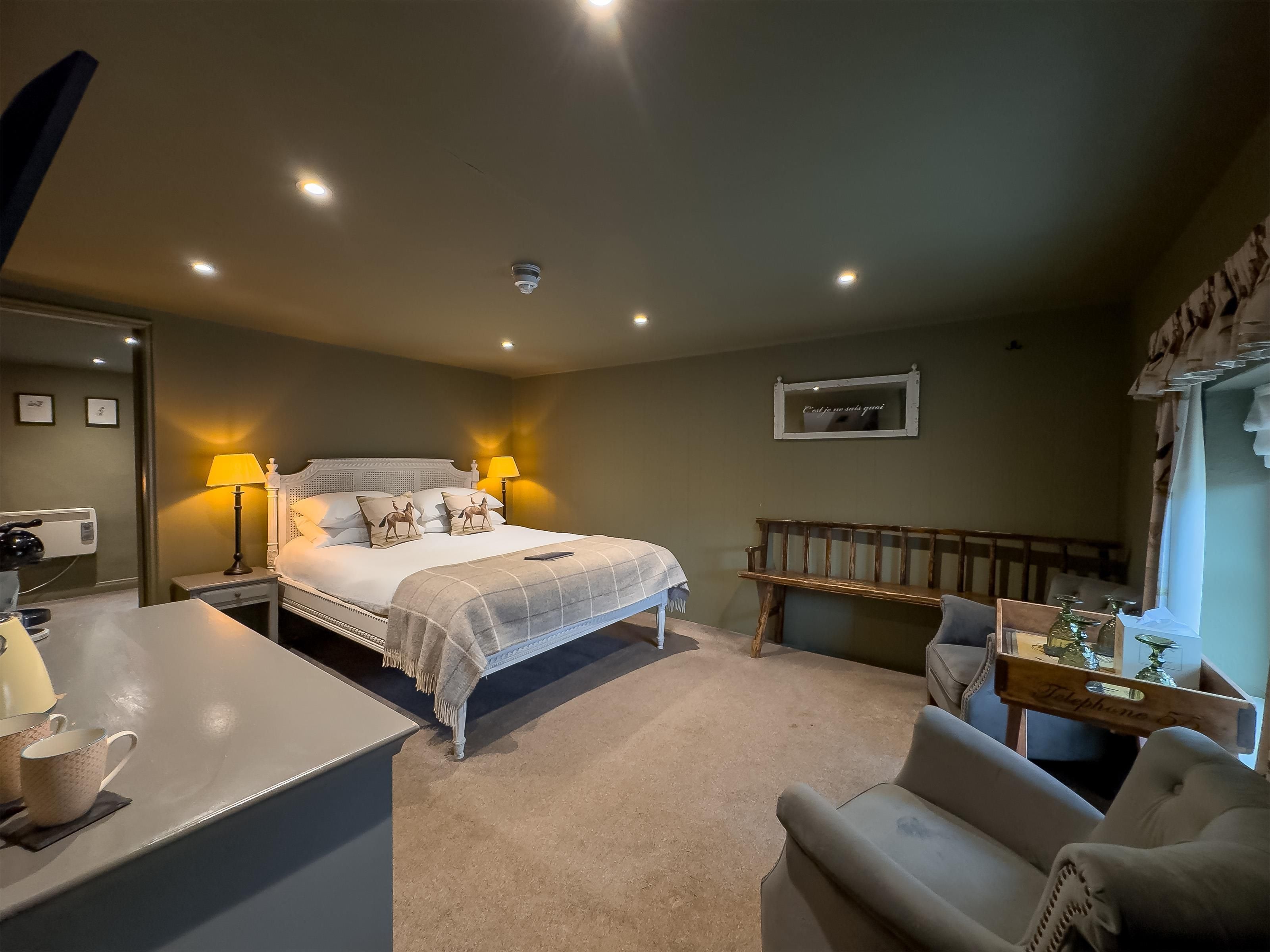 Habitación de hotel con cama, lámparas de noche, mesitas de noche, una silla, una mesita con bandeja, un espejo y un cuadro en la pared.