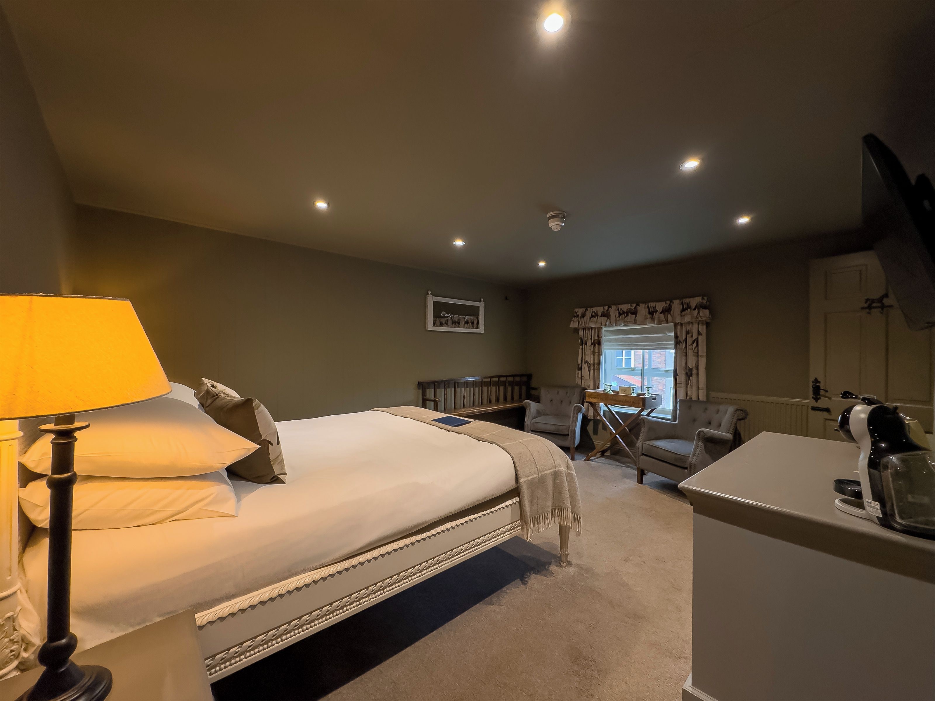 Une chambre d’hôtel avec un lit, un bureau, une chaise, une lampe et une fenêtre avec des rideaux.