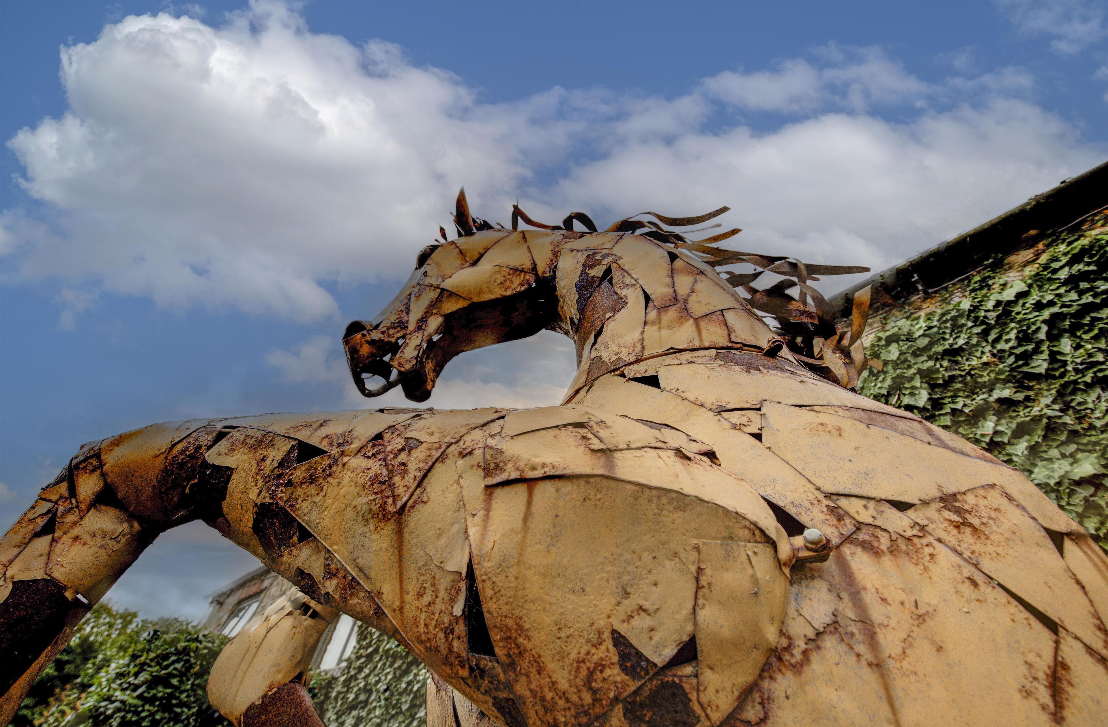 A large, rusted metal sculpture of a horse with a damaged head and missing parts, set against a backdrop of a partly cloudy sky and greenery.