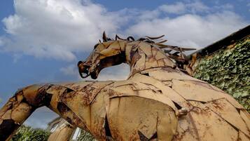 Una gran escultura de metal oxidado de un caballo con la cabeza dañada y partes faltantes, sobre un fondo de cielo parcialmente nublado y vegetación.