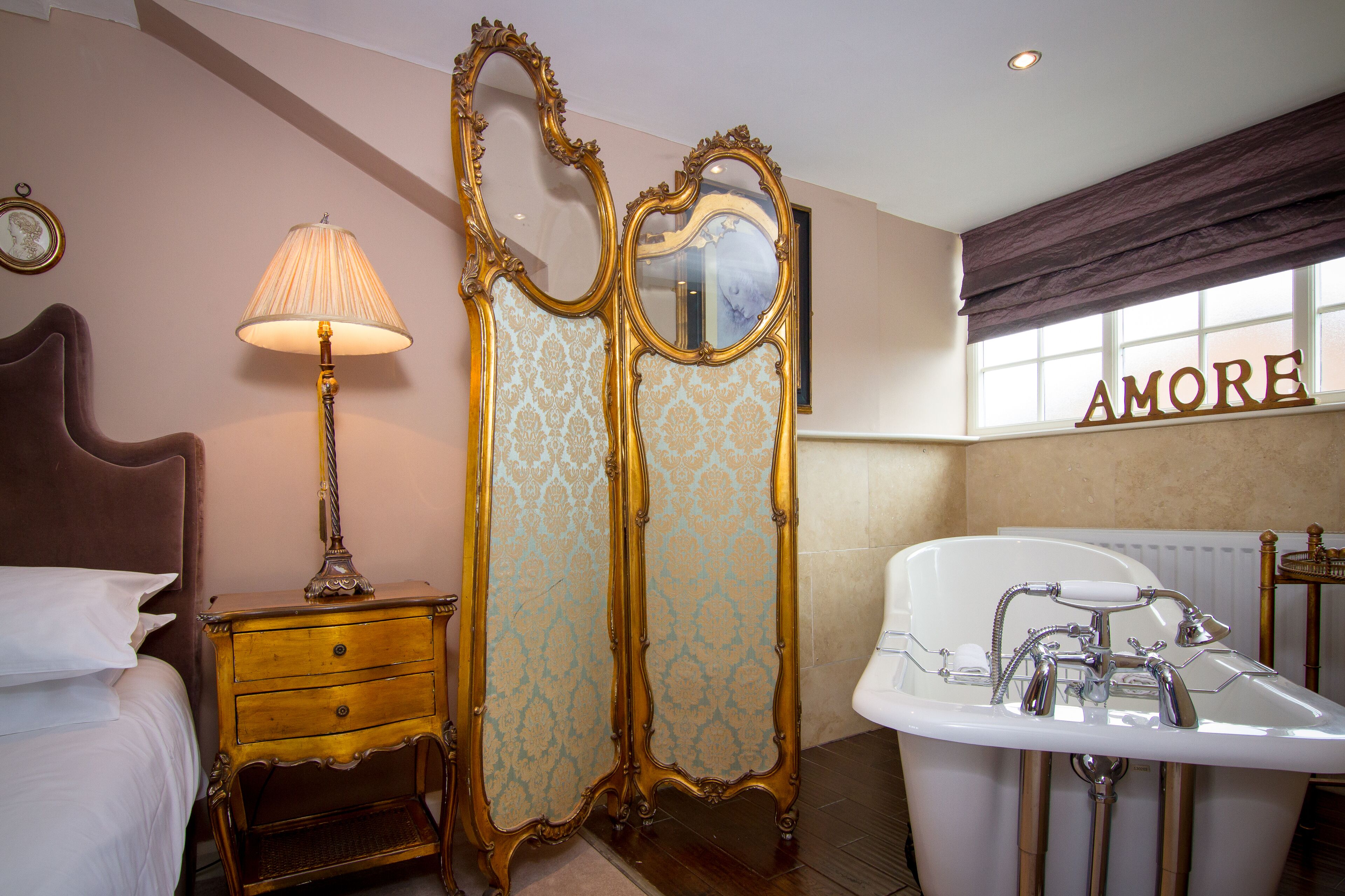 A bedroom with a bed, a nightstand with a lamp, a dressing table with a mirror, and a bathroom with a bathtub and sink.