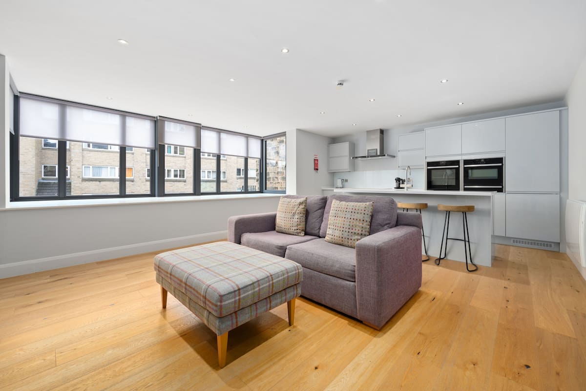 A modern apartment with a grey sofa, a plaid ottoman, and a kitchen with white cabinets and two ovens.