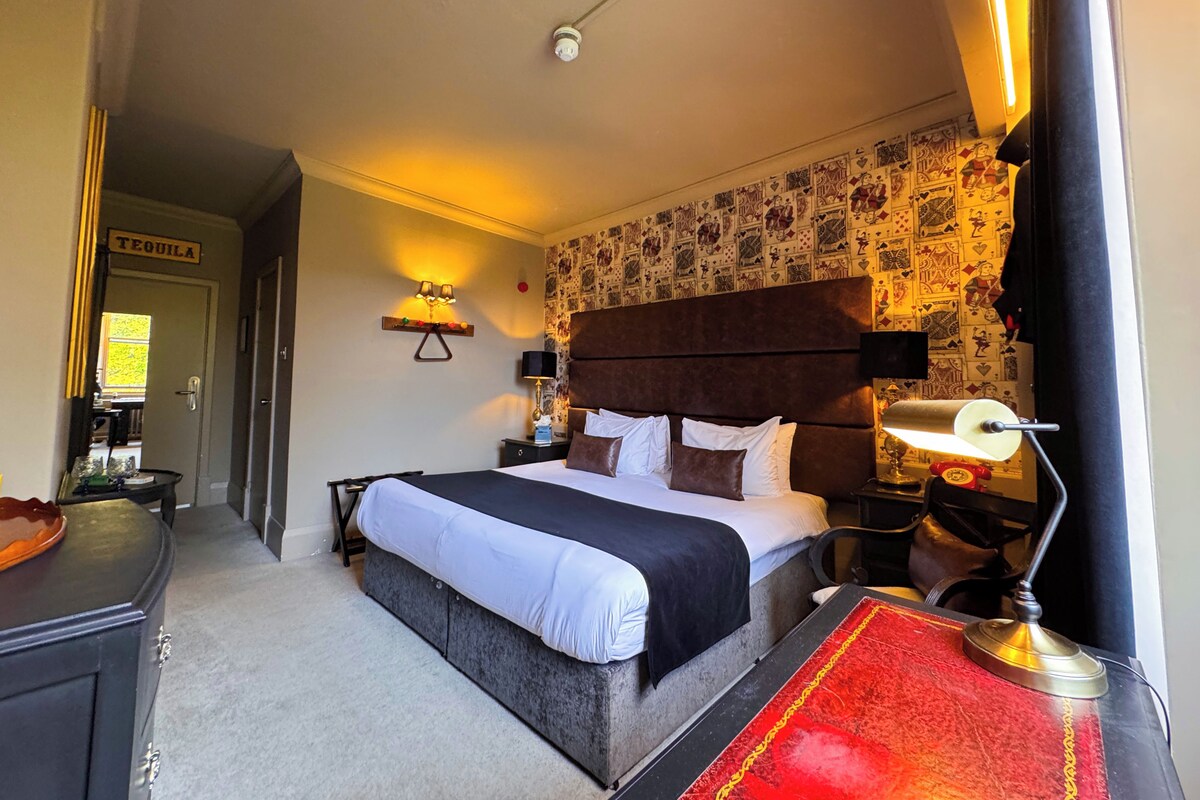 Habitación de hotel con cama, mesitas de noche, una lámpara de pared, alfombra roja y una puerta con la etiqueta 'TEQUILA'.