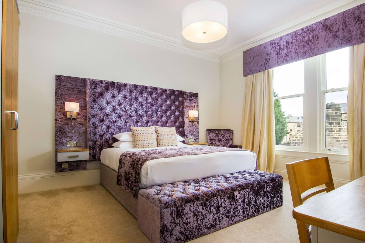 A bedroom with a large bed, a bedside table, a chair, and a window with curtains.