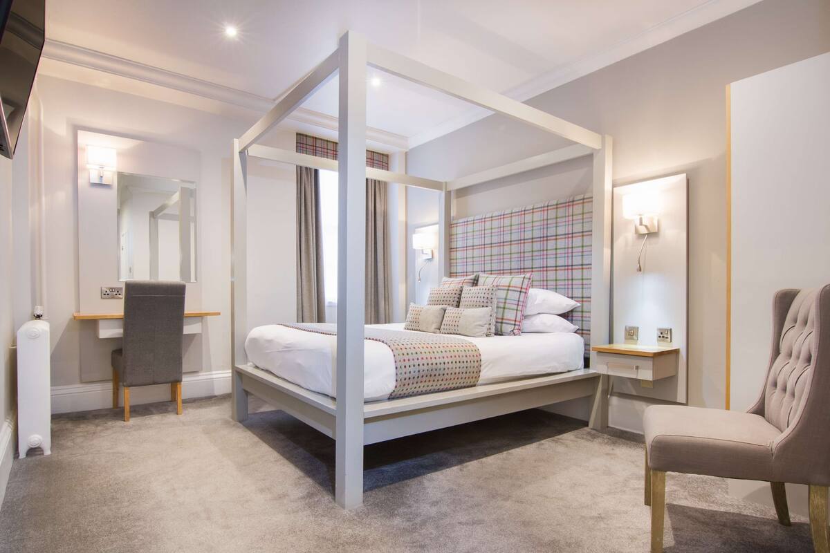 A four-poster bed with a white and gray color scheme, a desk with a chair, and a bedside table with a lamp.