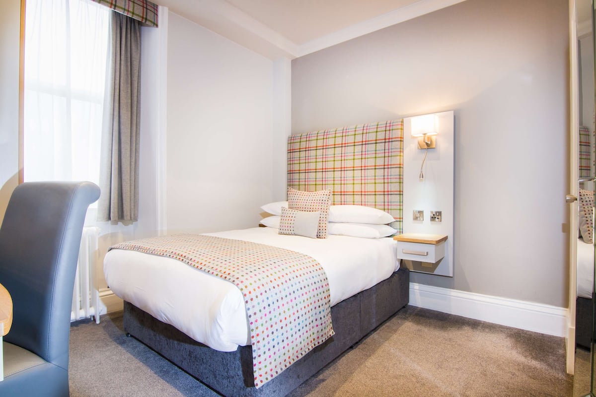 A hotel room with a bed, a chair, a nightstand, and a wall-mounted lamp.
