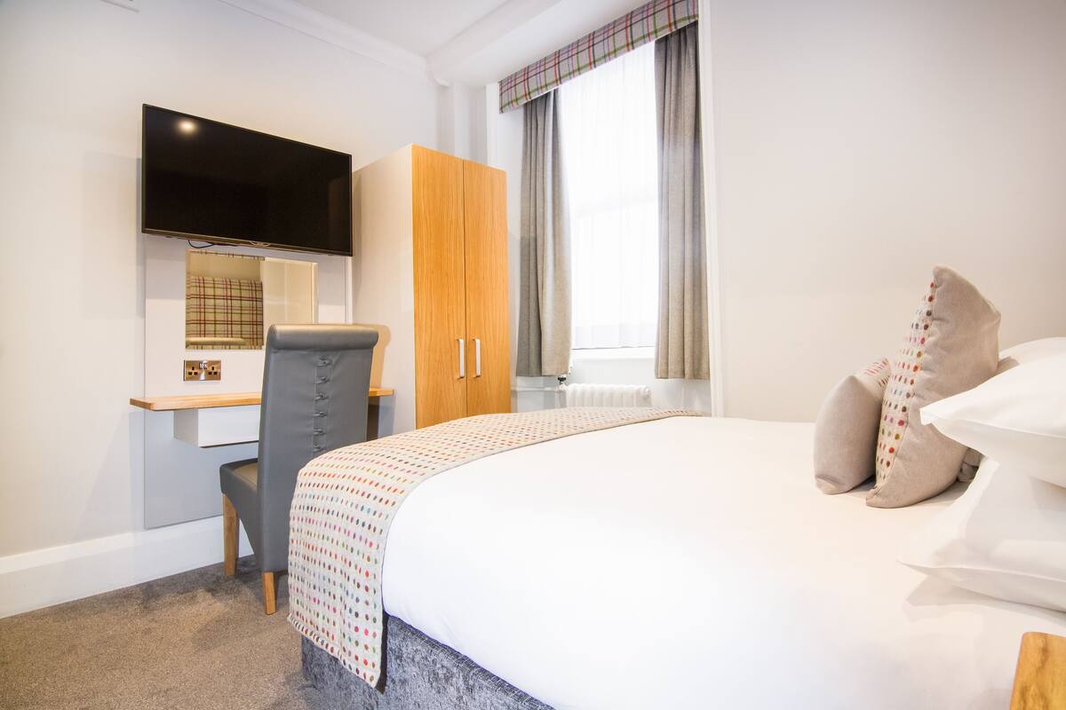 Quarto de hotel com uma cama, uma escrivaninha com cadeira, televisão fixada na parede e janela com cortinas.