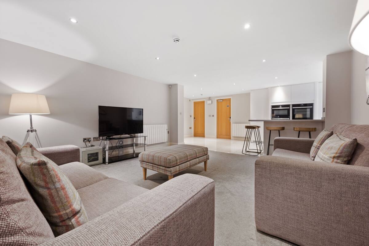 A modern living room with a flat-screen TV, a sofa, an ottoman, and a lamp.