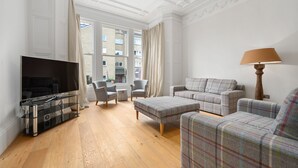 A living room with a flat-screen TV, two grey armchairs, a plaid sofa, a wooden floor, and a window with curtains.