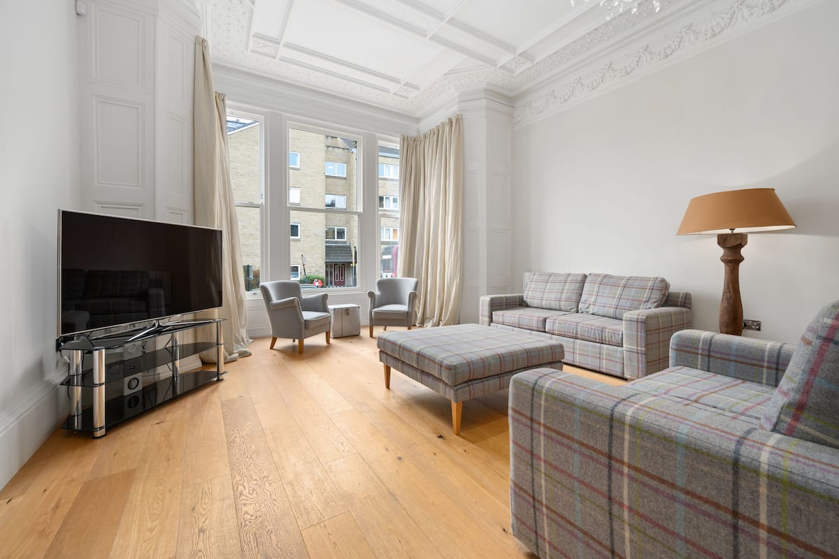 A living room with a flat-screen TV, two grey armchairs, a plaid sofa, a wooden floor, and a window with curtains.