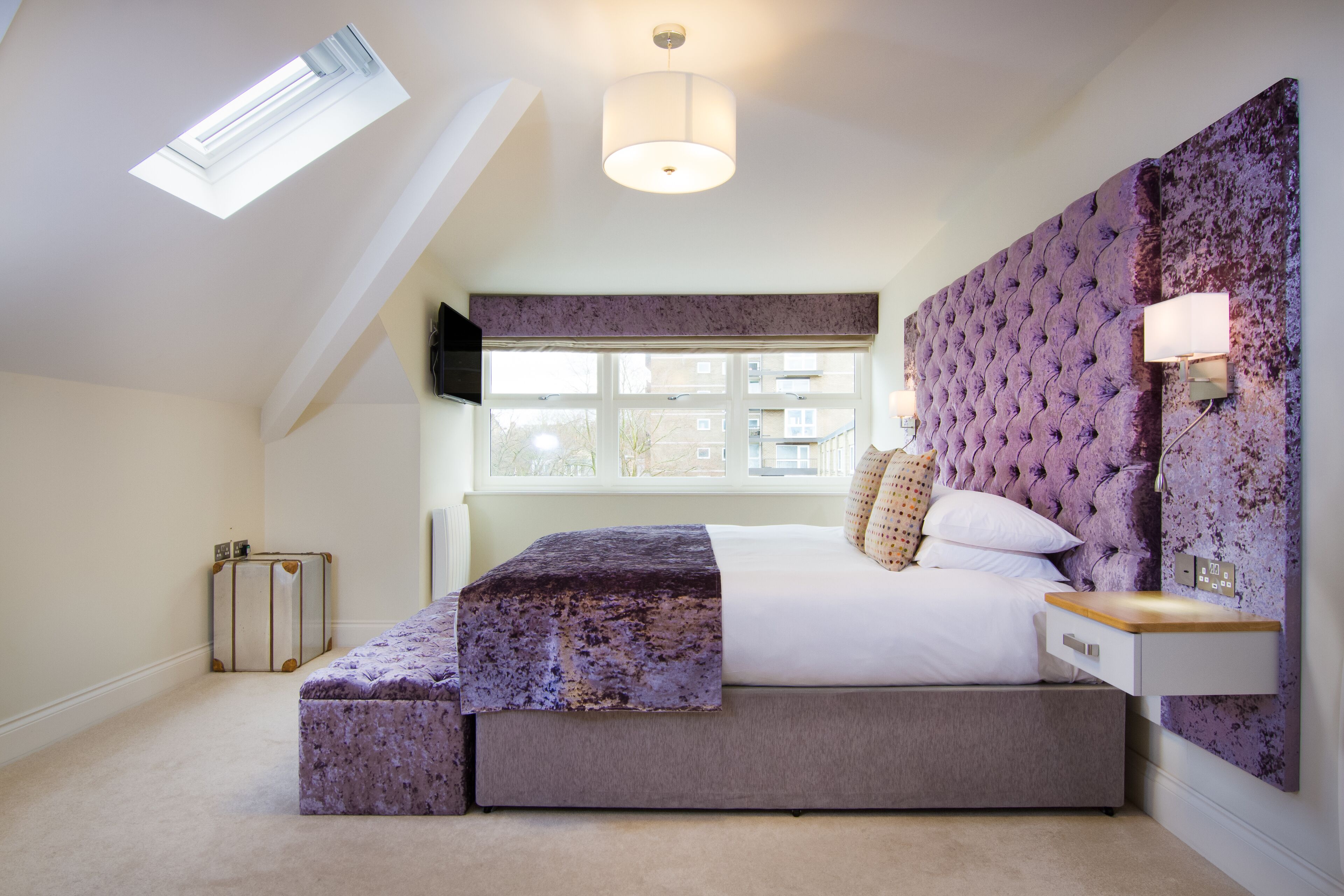 A bedroom with a large bed, a bench, a window, and a ceiling light.