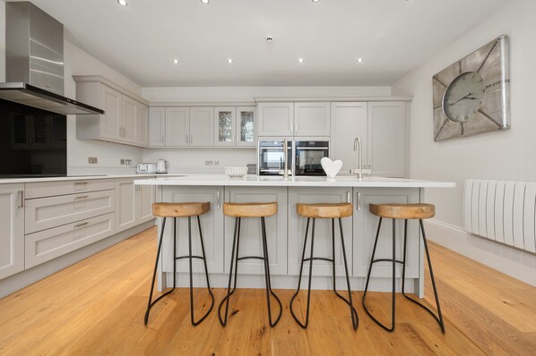 A modern kitchen with white cabinets, a central island with stools, stainless steel appliances, and a wall clock. -  White Hart Hotel & Apartments (Harrogate)