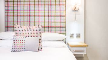 A neatly made bed with white linens and colorful polka dot pillows, a plaid-patterned headboard, and a bedside table with a lamp.