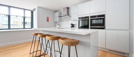 Cocina moderna con armarios blancos, una isla central con taburetes y un amplio ventanal que proporciona luz natural.