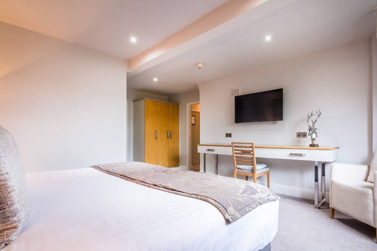 Quarto de hotel com uma cama grande, uma mesa de trabalho, uma cadeira e uma televisão na parede.