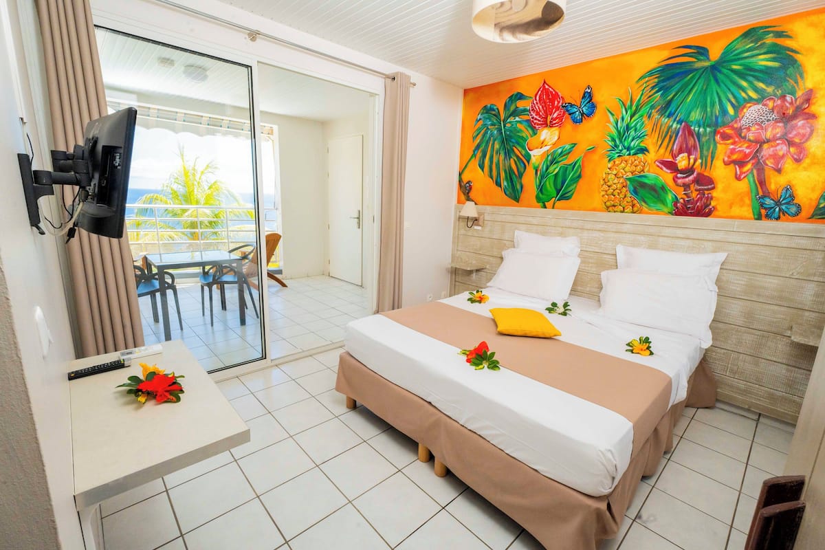 A hotel room with a bed, a TV, a desk with a flower, and a mural of tropical flowers.