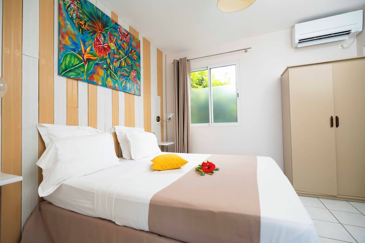 A hotel room with a bed, a painting on the wall, an air conditioning unit, and a wardrobe.