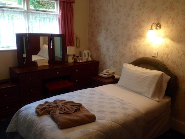 A bedroom with a bed, a dresser, a mirror, a nightstand, a lamp, and a wall-mounted light. - Grange Lodge Hotel (St. Peter Port)