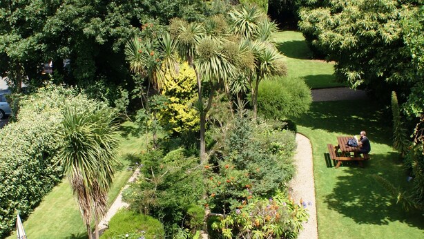 A garden with a circular wooden table and benches, surrounded by various plants and trees. - Grange Lodge Hotel (St. Peter Port)