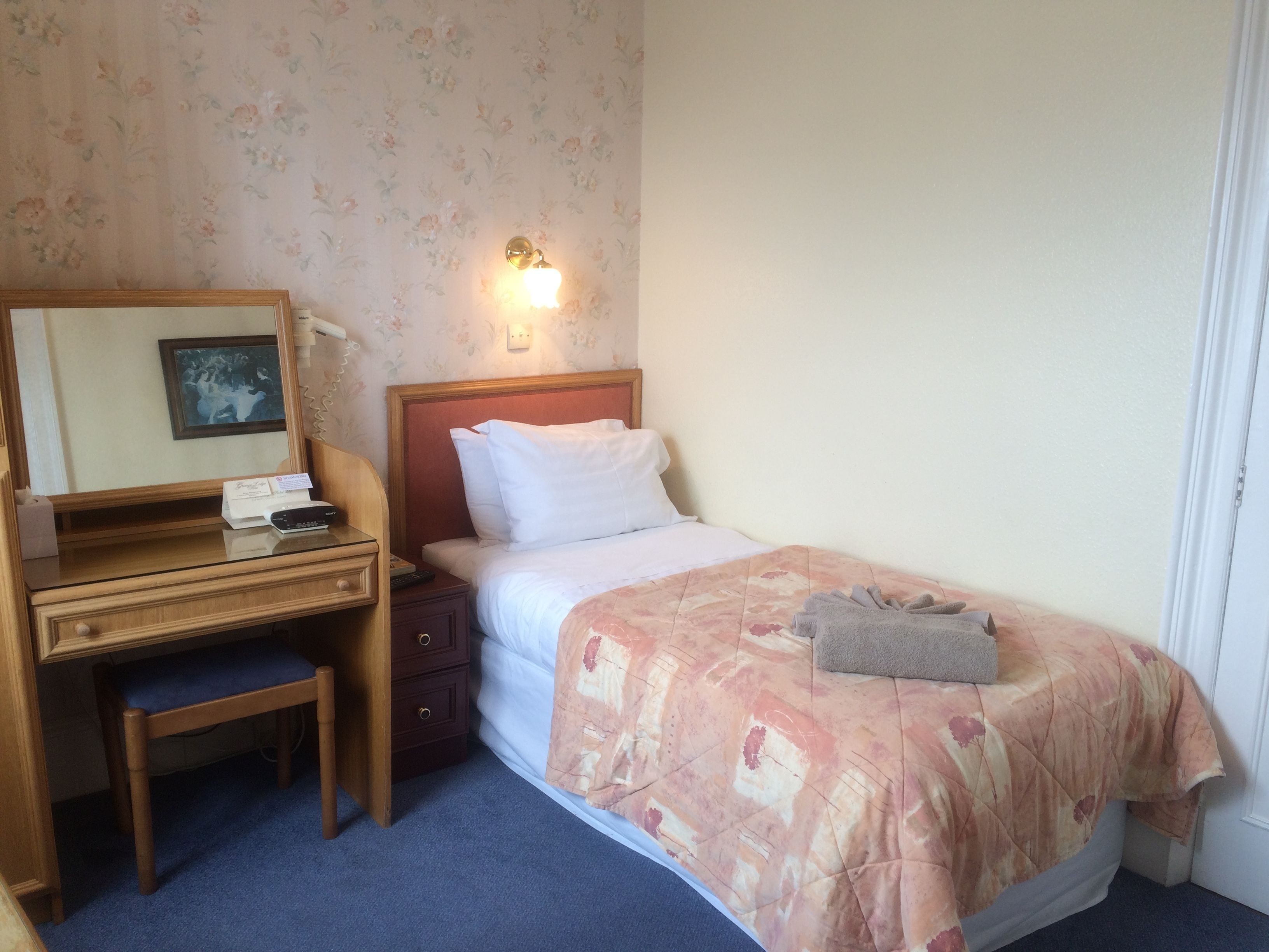 Una habitación de hotel con un escritorio de madera, una cama con colcha estampada, una mesita de noche y una lámpara de pared.