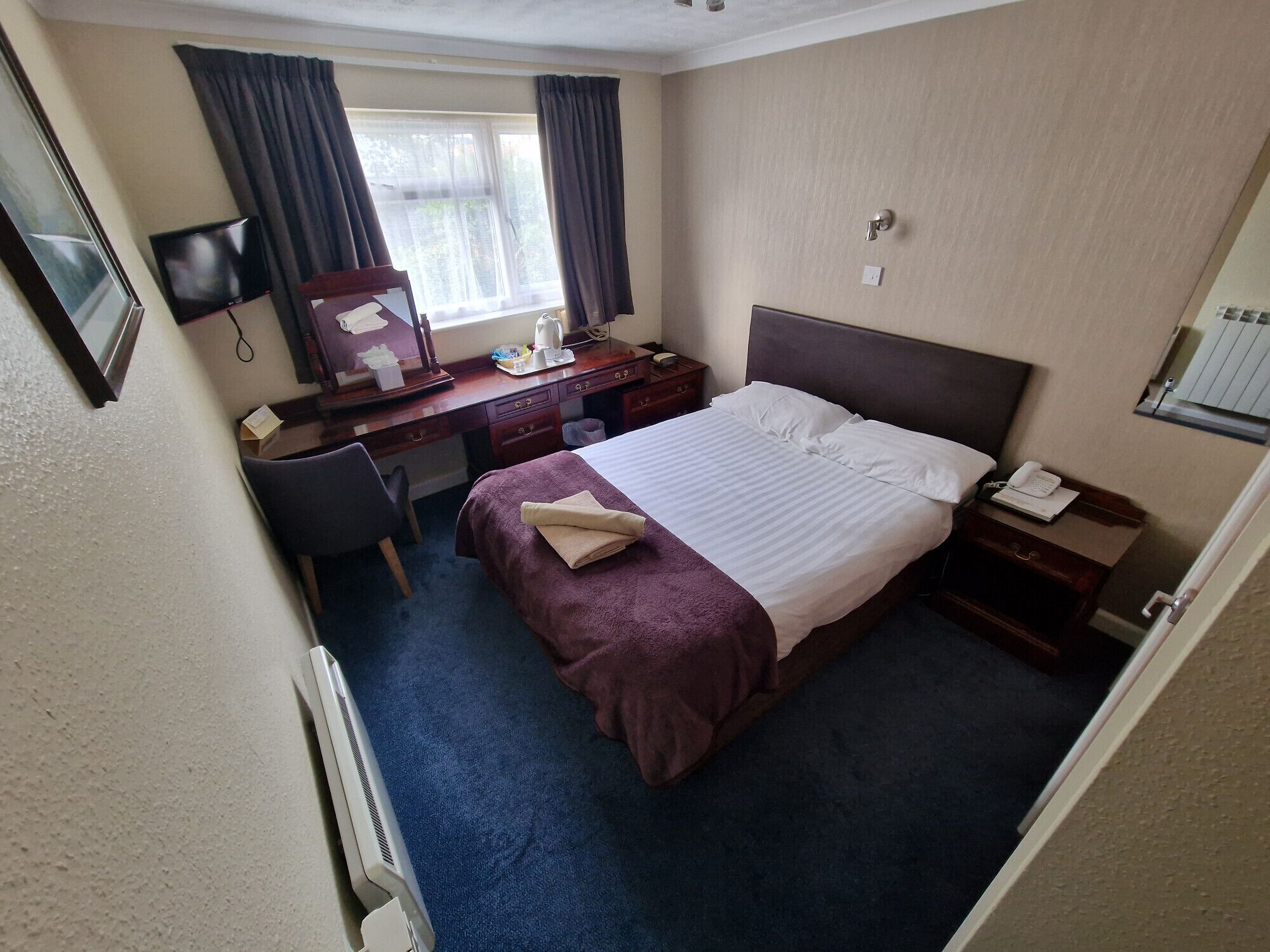 A hotel room with a bed, a desk, a chair, a TV, a window with curtains, and a bedside table with a phone.