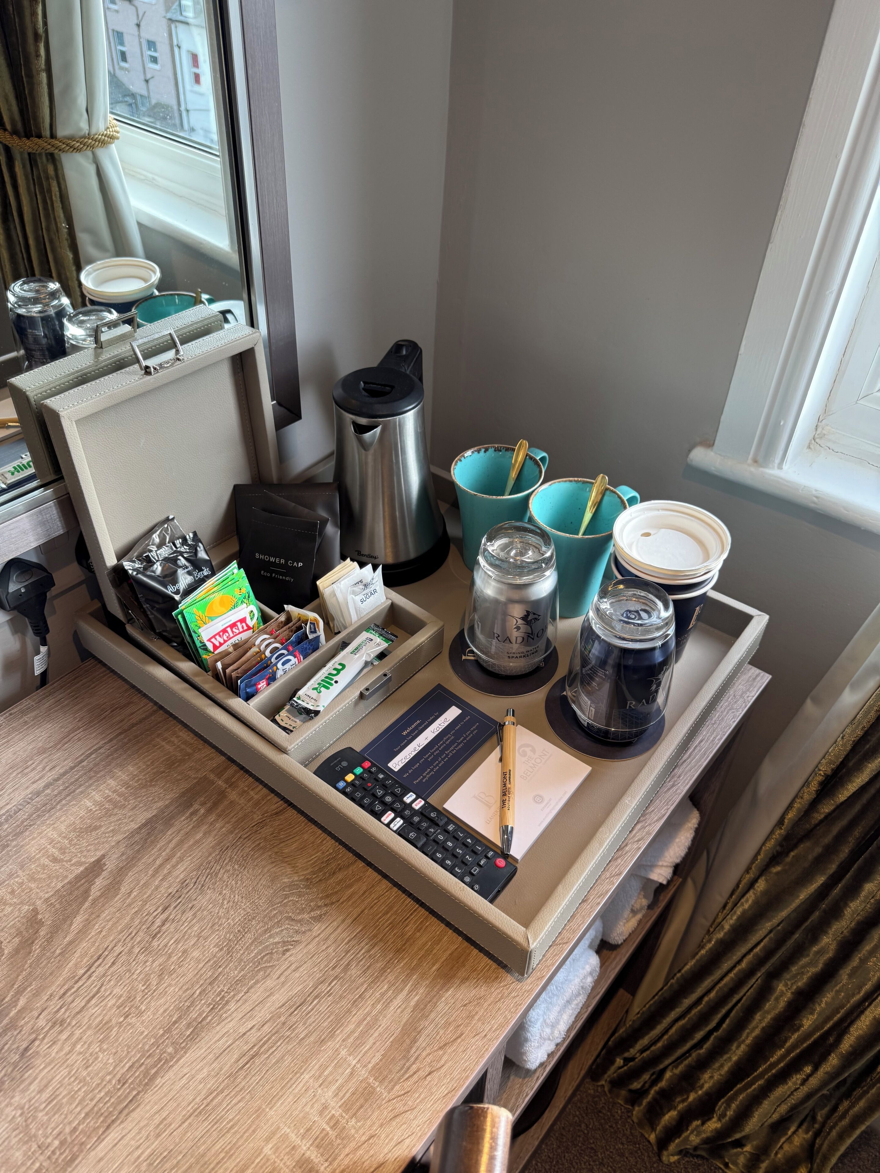 A neatly organized desk with a laptop, a coffee maker, a mug, a cup, a tray with snacks, a pen, and a notepad.
