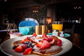 Desayuno con tortitas cubiertas de fresas, arándanos y frambuesas, acompañado de un vaso de zumo de naranja y salsas.
