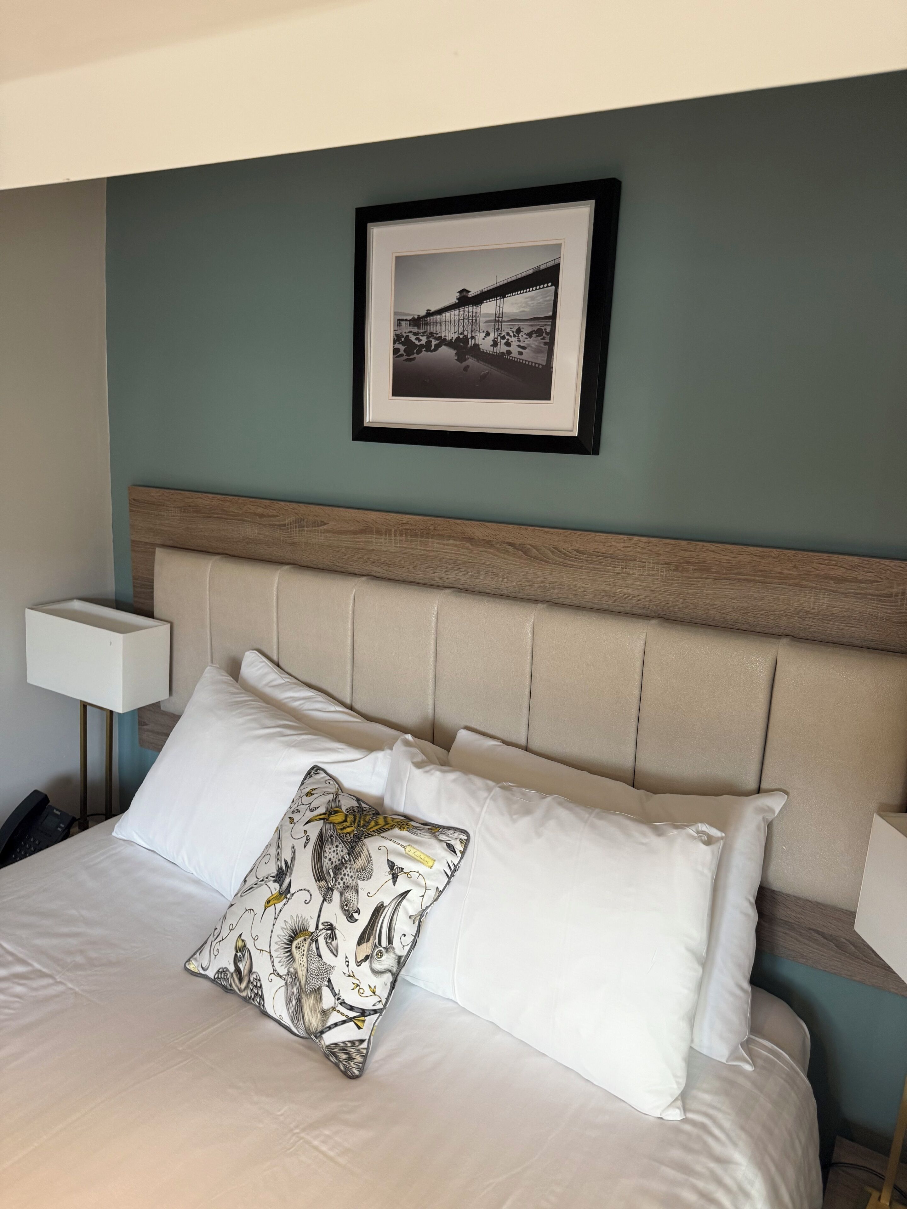 Een keurig opgemaakt bed met witte lakens en een decoratief kussen, een houten hoofdbord, een bedlampje en een lijst met een foto van een brug aan de muur.