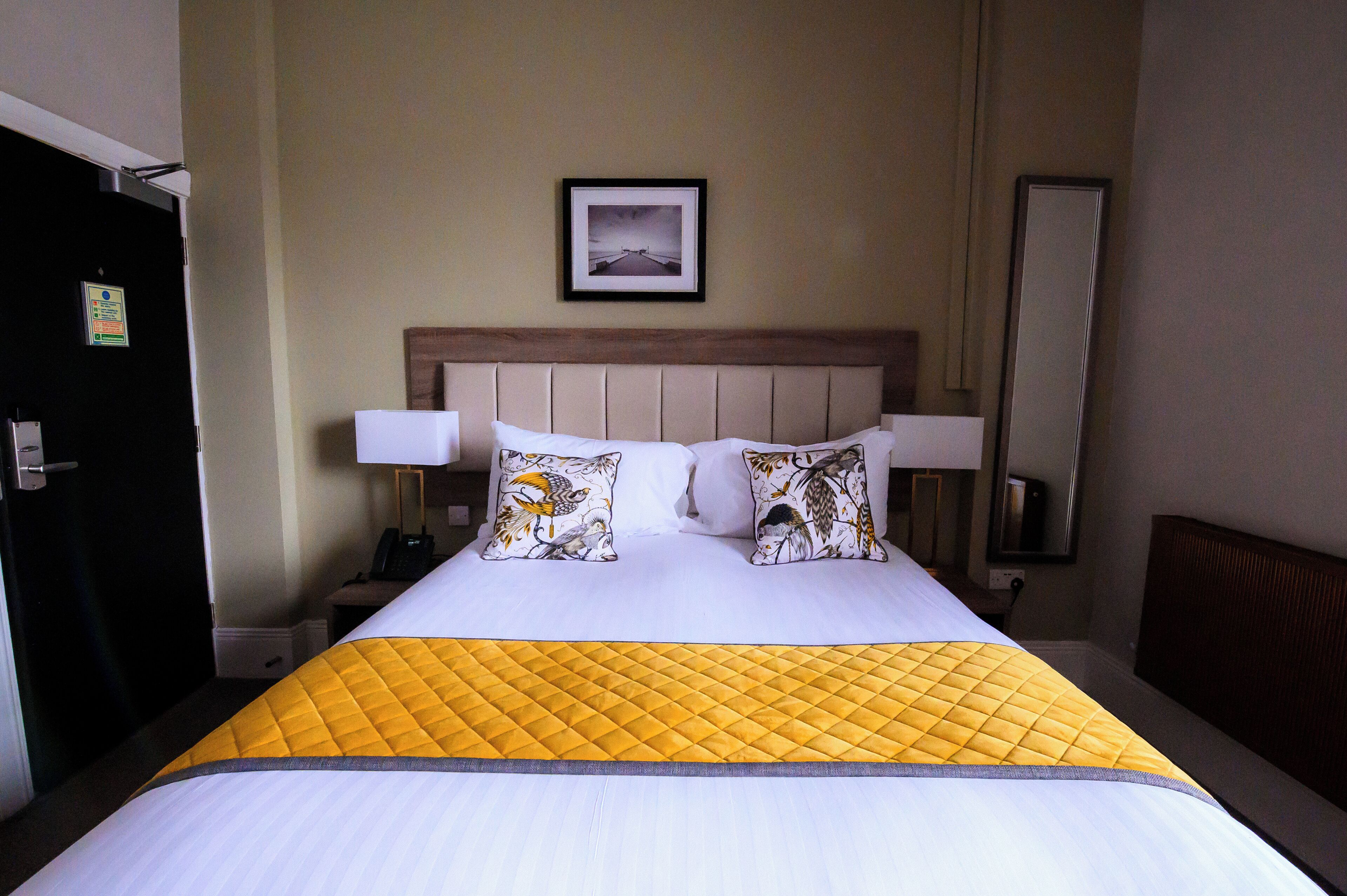 A hotel room with a bed, a nightstand, a lamp, a framed picture on the wall, and a door.