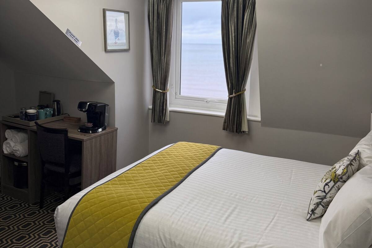 Habitación de hotel con cama, escritorio, silla, cafetera y vista al océano a través de una ventana.