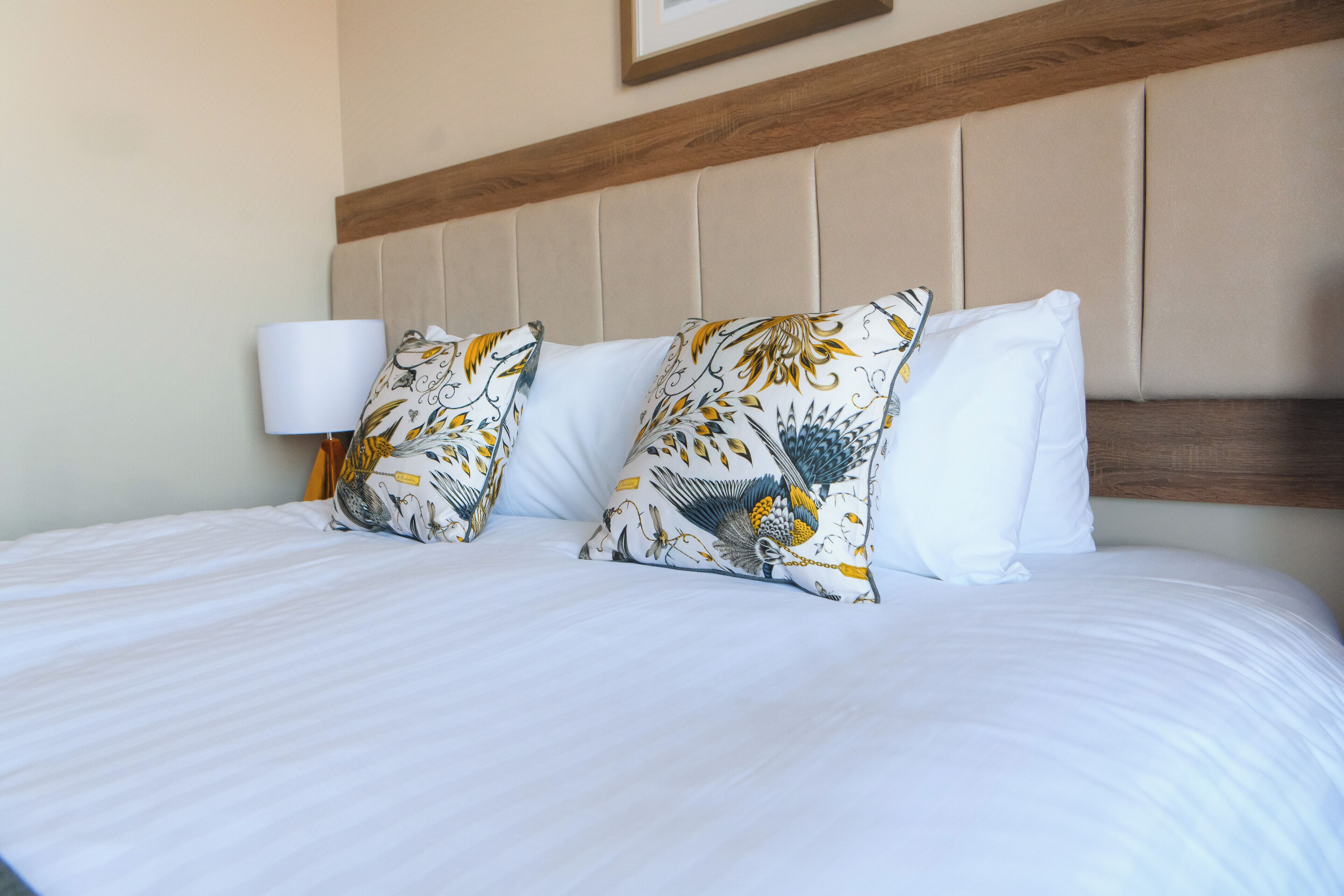 A neatly made bed with white linens and patterned pillows, a wooden headboard, and a wall-mounted lamp.