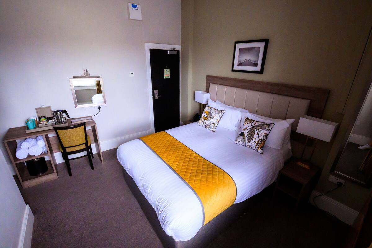 Habitación de hotel con cama, escritorio, silla, puerta y un cuadro colgado en la pared.