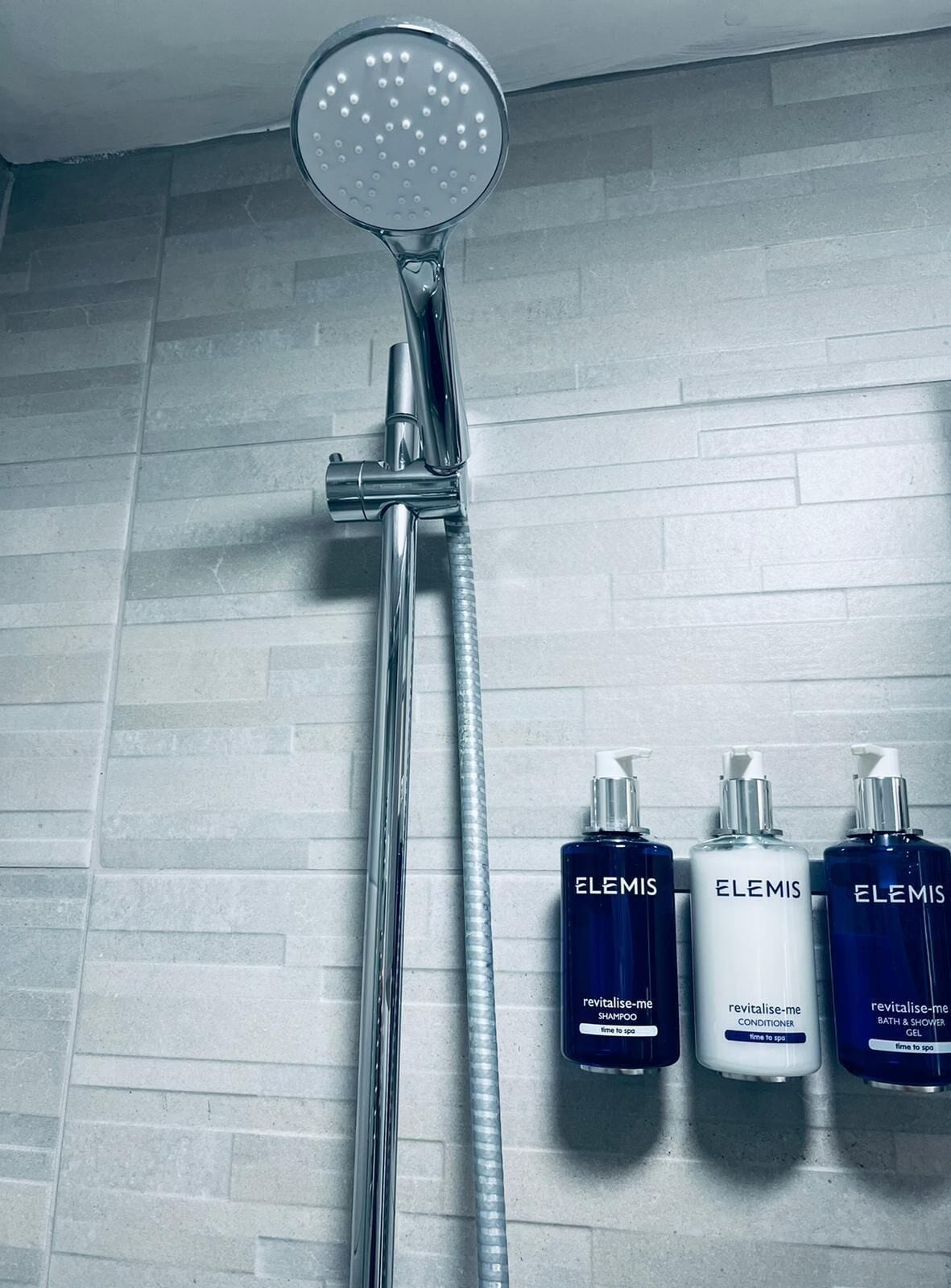 A shower with a chrome head and a flexible hose, and three bottles of Elemis skincare products.