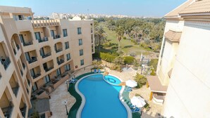 A swimming pool surrounded by a building with balconies, a view of a golf course, and a cityscape in the background.