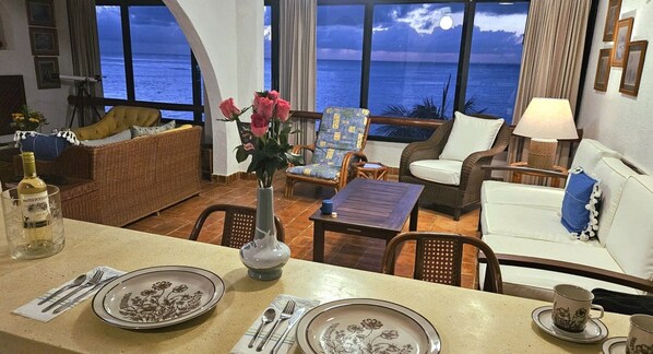 Una habitación con vista al océano, un comedor con platos y cubiertos, y mobiliario de mimbre.