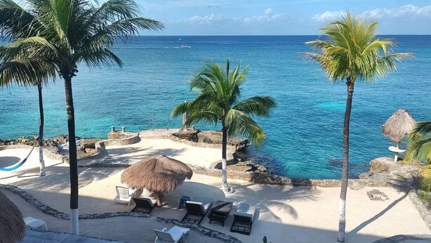 Un complejo turístico en la playa con palmeras, sillas de descanso, una cabaña de techo de paja y una piscina.