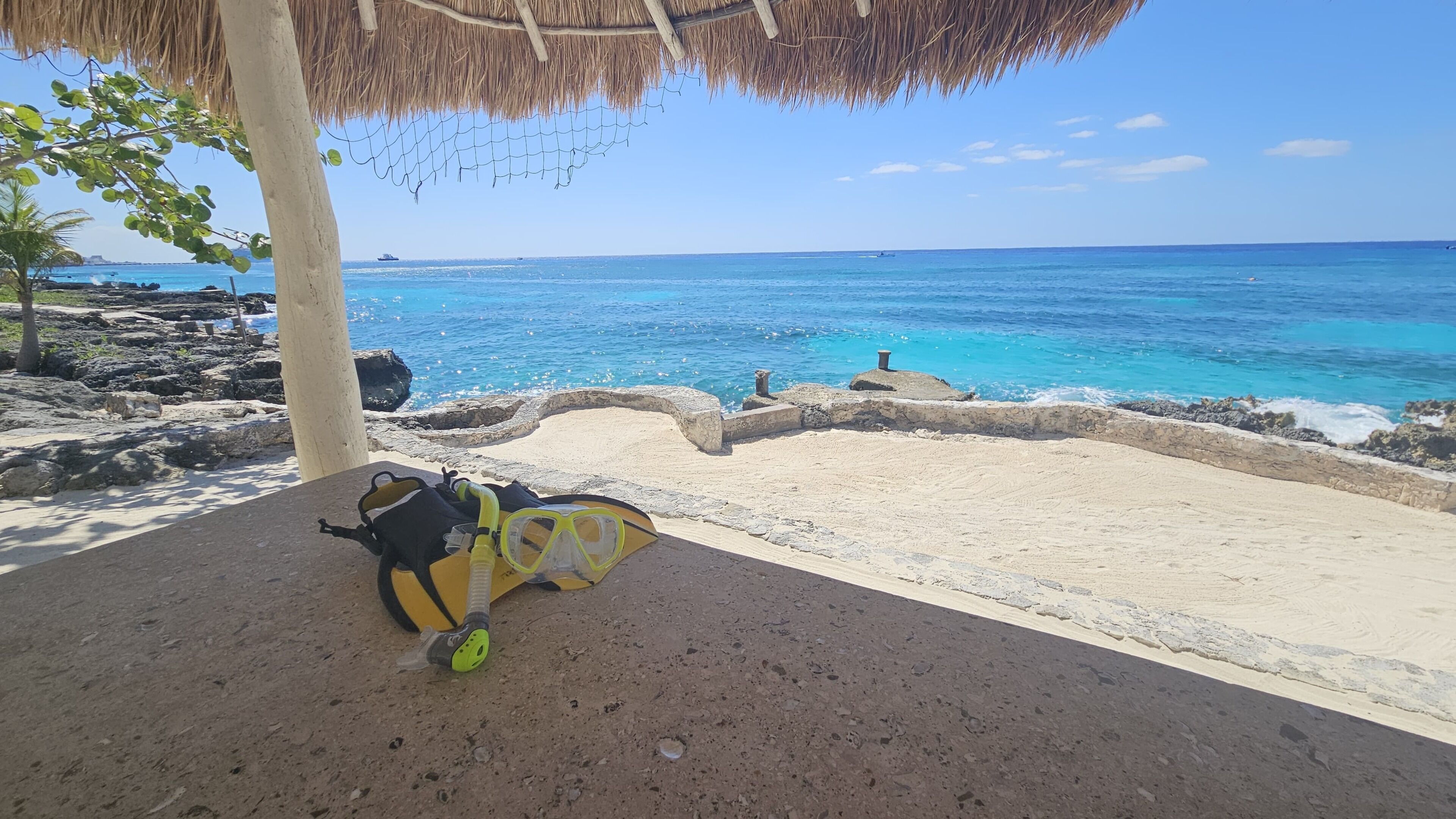 Una playa con una estructura de techo de paja, un snorkel y una máscara en el suelo, y el océano al fondo.