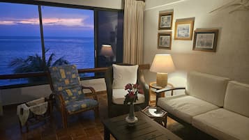 A room with a view of the ocean, a sofa, a chair, a table with a lamp, and framed pictures on the wall.