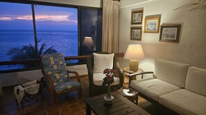 Een kamer met uitzicht op zee, een bank, een stoel, een tafel met een lamp en lijstjes met foto's aan de muur.