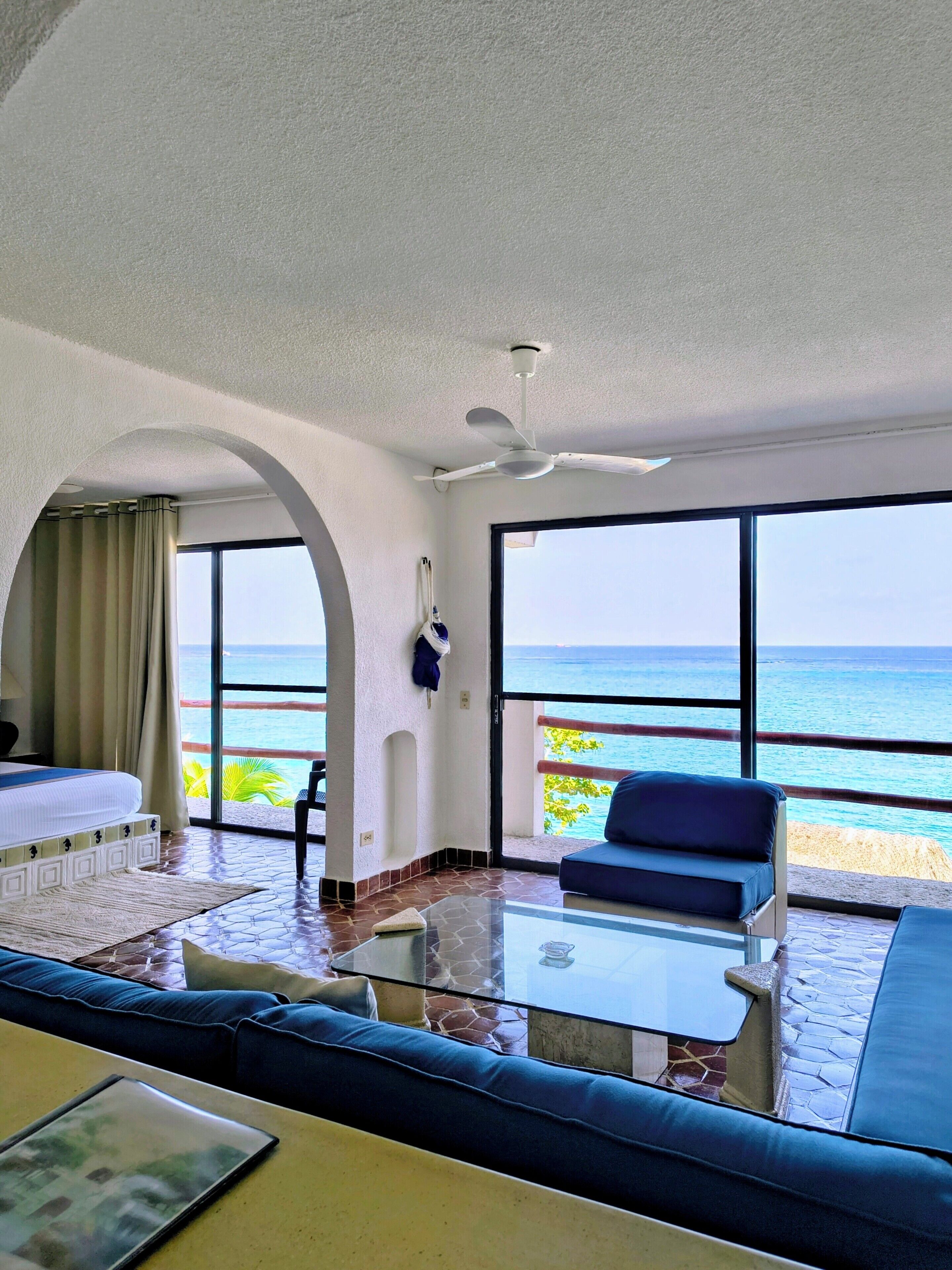 A hotel room with a balcony, a bed, a sofa, a table, and a view of the ocean.