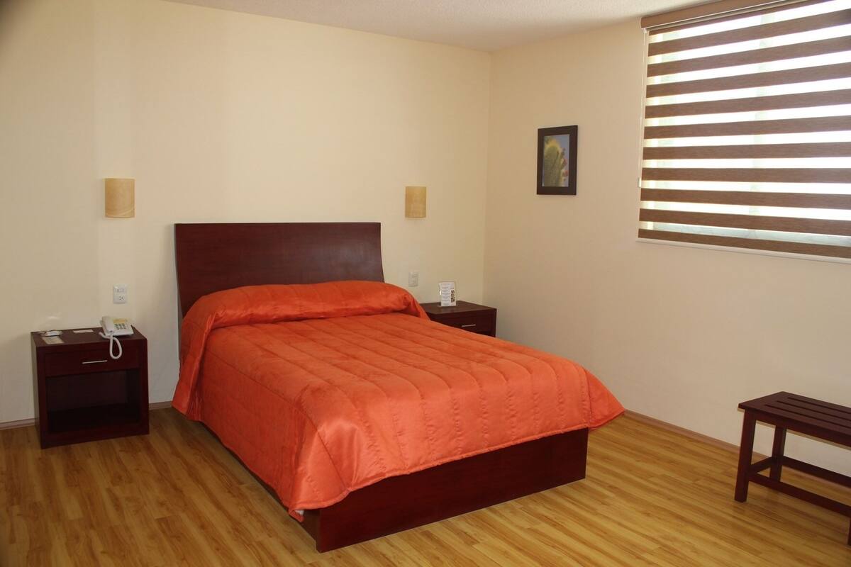 Une chambre d’hôtel avec un lit en bois, deux tables de chevet, une petite table d’appoint, un petit banc en bois et une fenêtre avec des stores.